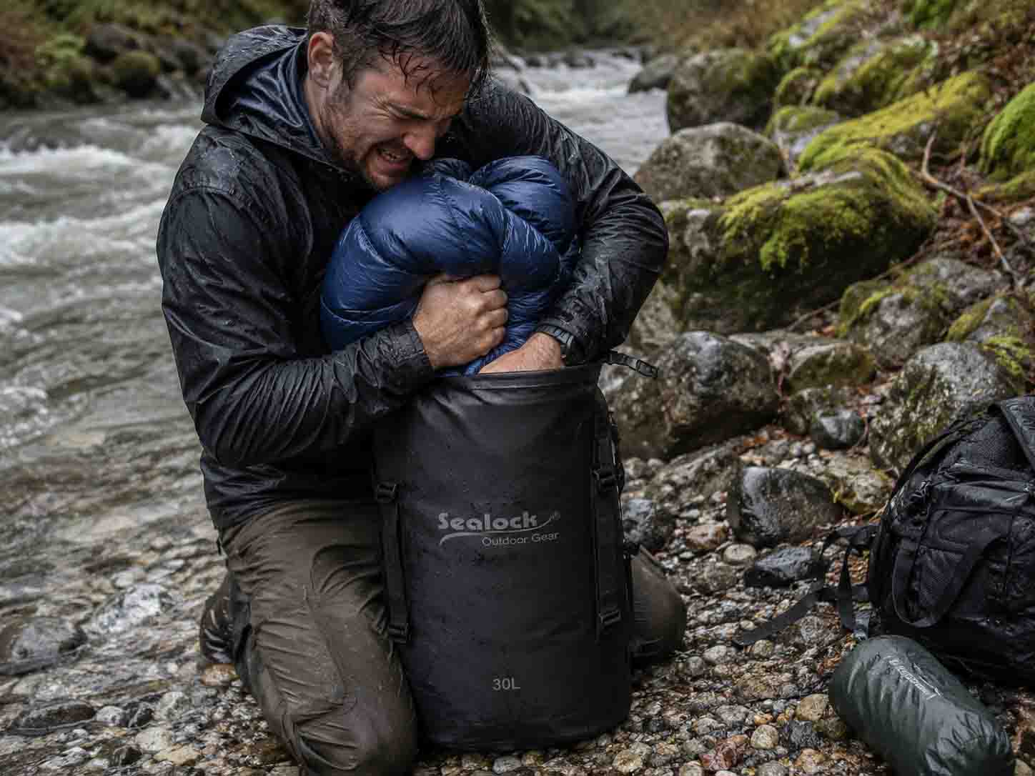 Real gear test failure photo of a hiker struggling to compress an uncompressed synthetic sleeping bag inside a stiff 30L matte TPU dry bag on river rocks Real gear test failure photo of a hiker struggling to compress an uncompressed synthetic sleeping bag inside a stiff 30L matte TPU dry bag on river rocks
